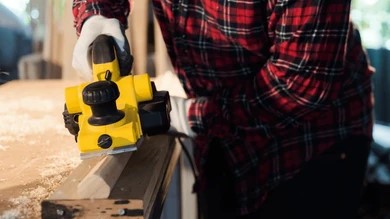 person using yellow electric hand planer on wood board with shavings