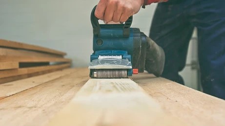 close up view of a blue belt sander being guided along a narrow piece of lumber