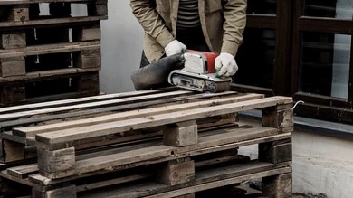 person using a power belt sander to smooth a wooden plank atop stacked pallets