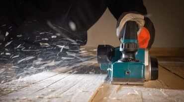 a worker using a turquoise electric hand planer to smooth a wooden surface while shavings fly