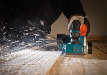 a worker using a turquoise electric hand planer to smooth a wooden surface while shavings fly